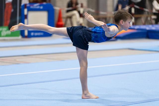 Young gymnast performs an arabesque balance with extended arms during floor exercise routine on blue training mat