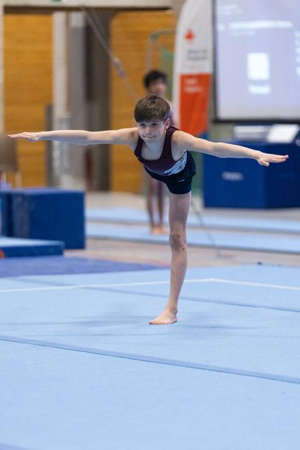 Young gymnast performs arabesque balance on floor mat, arms extended horizontally, demonstrating focus and control in gym