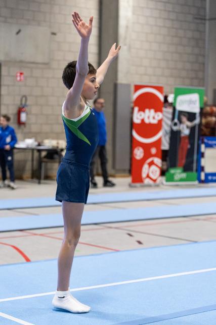 Young gymnast in blue and green leotard stands with arms raised overhead during floor routine at indoor gymnasium