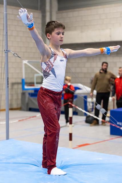 Young gymnast in white and burgundy uniform strikes an artistic pose with arms extended on blue competition mat