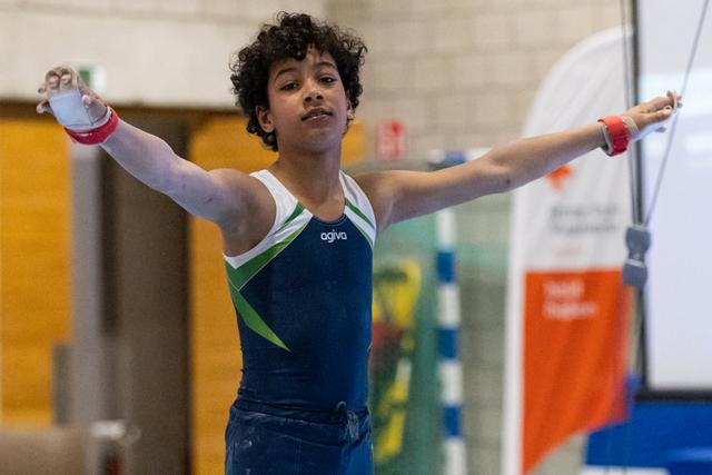 Young gymnast performs on rings with arms extended, showing concentration and control in blue and green leotard