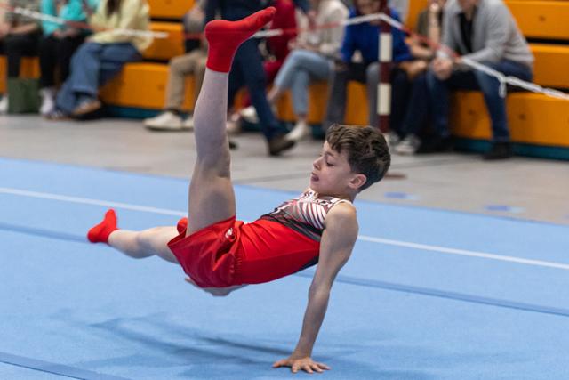 Young gymnast performs dynamic breakdancing freeze move on blue floor mat with spectators watching from bleachers