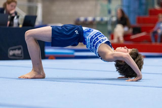 Young gymnast performs a bridge pose during floor routine, demonstrating flexibility and strength on blue competition mat