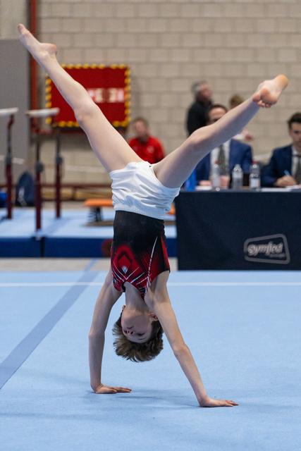 Young gymnast performs a cartwheel during floor exercise, legs extended in split position, with judges seated at table behind