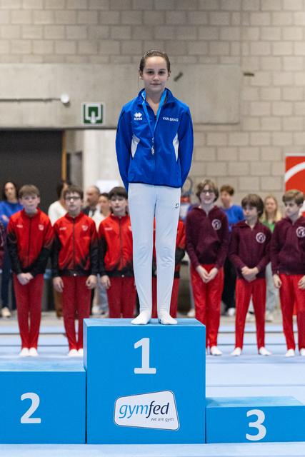 Young gymnast in blue tracksuit stands proudly on first place podium at gymfed event with teammates watching behind