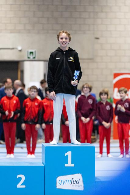 Young gymnast in black tracksuit stands on first place podium holding trophy, beaming with pride as teammates watch