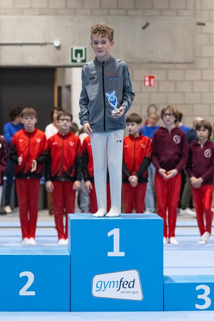Young gymnast stands proudly on first place podium holding trophy, with teammates in red tracksuits watching from behind