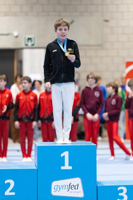 Young gymnast stands on first place podium holding gold medal, smiling proudly while teammates watch from background