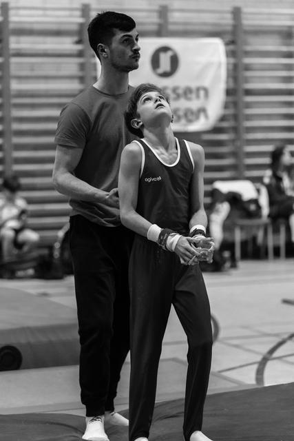 Young gymnast and coach stand side by side, both gazing upward with focused expressions during training at an indoor gym