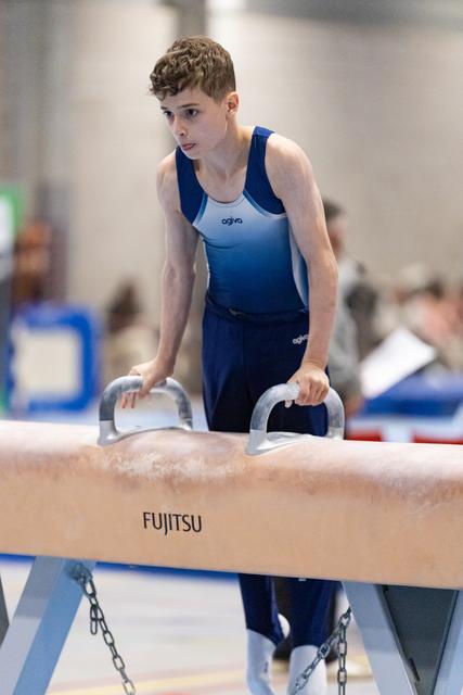 Young gymnast grips pommel horse handles with focused expression, preparing for routine in blue competition uniform