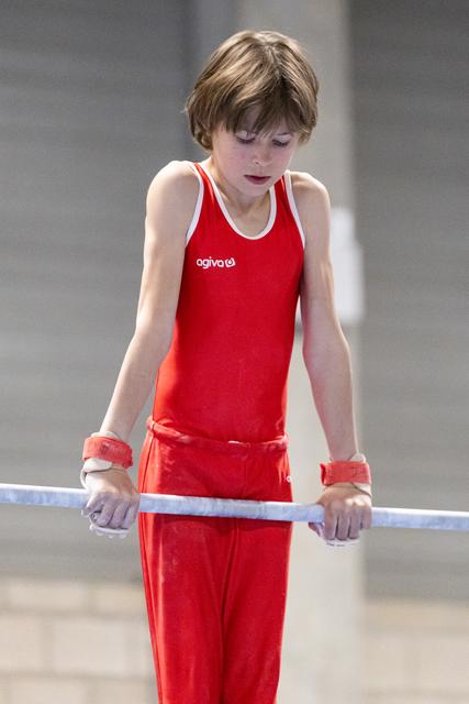 Young gymnast in red uniform focuses intently on the parallel bars, hands gripped and eyes downcast in concentration
