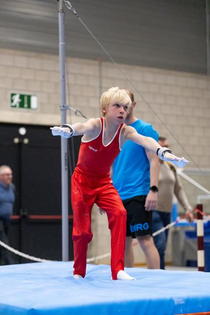 Young gymnast in red uniform dismounts from parallel bars with intense focus, coach observing in background