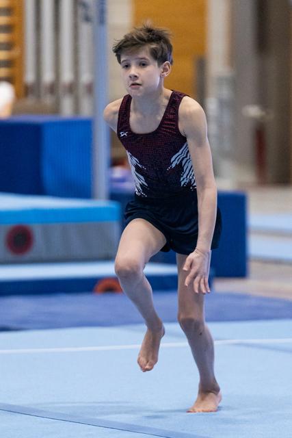 Young gymnast in black leotard concentrates during floor routine landing, barefoot on blue mat in training facility