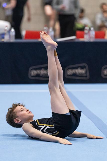 Young gymnast performs floor routine with legs extended vertically, demonstrating flexibility and balance on blue mat