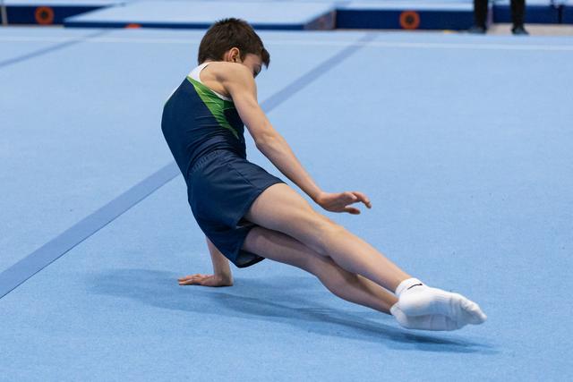Young gymnast performs floor routine with extended leg stretch on blue mat, showing strength and flexibility