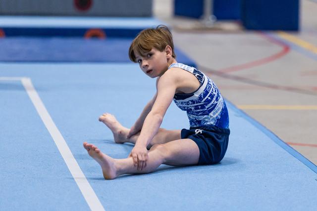 Young gymnast in blue sparkly leotard performs stretching position on floor mat with focused expression