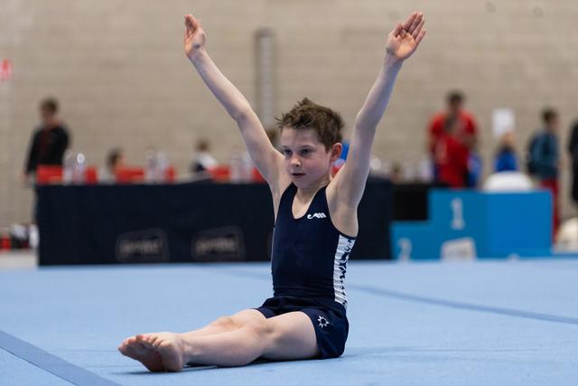 Young gymnast in black and white leotard sits on floor with arms raised overhead, displaying focused expression during routine