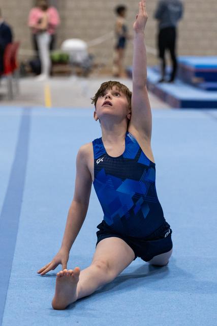 Young gymnast in blue leotard strikes an artistic floor routine pose, one arm raised gracefully while kneeling on blue mat