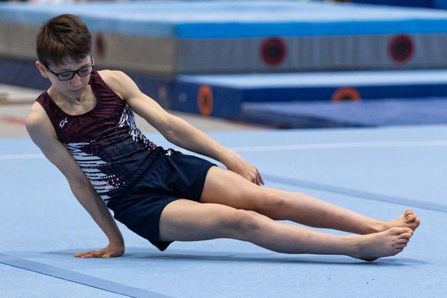 Young gymnast in glasses performs seated floor exercise move, legs extended forward in athletic pose on blue mat
