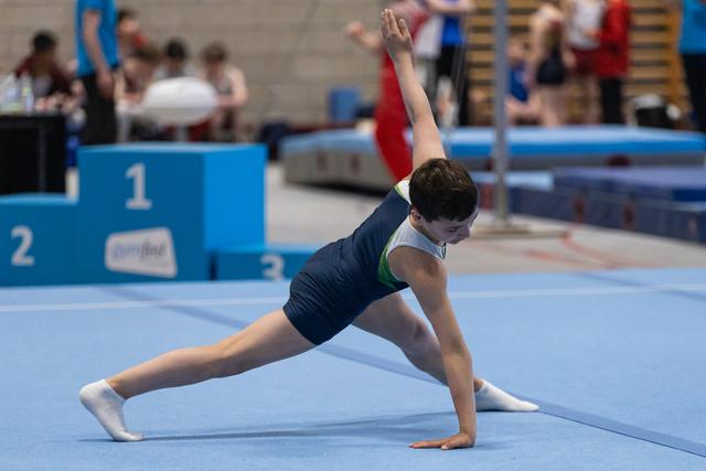 Gymnast performs a side split pose during floor routine, one arm extended upward, in competition hall with podiums visible