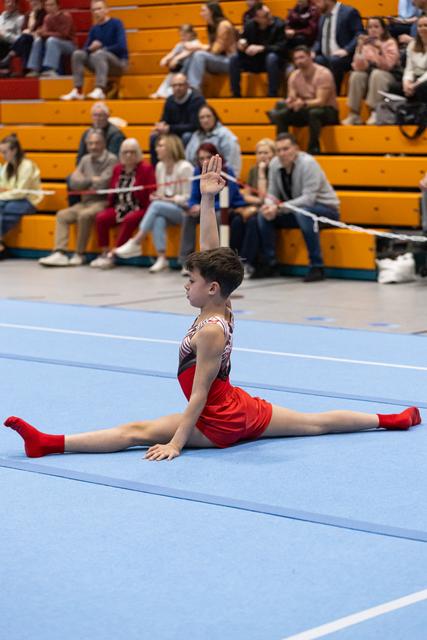 Young gymnast in red performs split on blue floor mat with one arm raised as audience watches from wooden bleachers