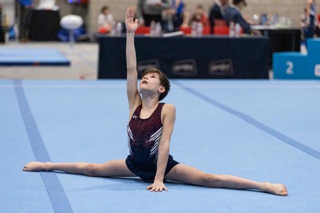 Young gymnast performs a floor split with one arm raised, demonstrating flexibility and poise on the blue competition mat.