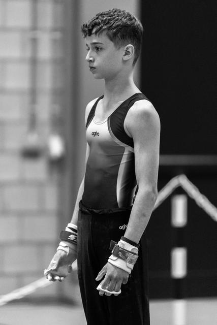 Young gymnast stands in profile, wearing black and white leotard with hand grips, displaying intense focus before his routine