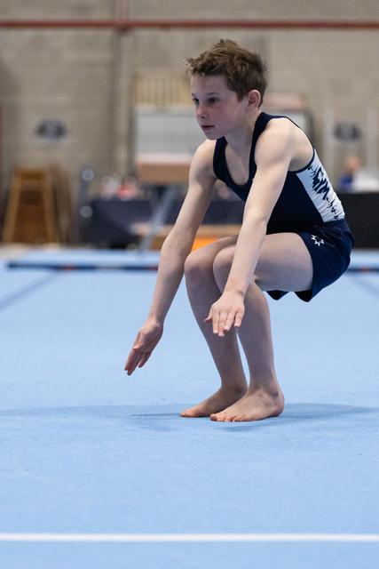 Young gymnast in navy leotard crouches in focused preparation on blue floor mat, arms extended in starting position