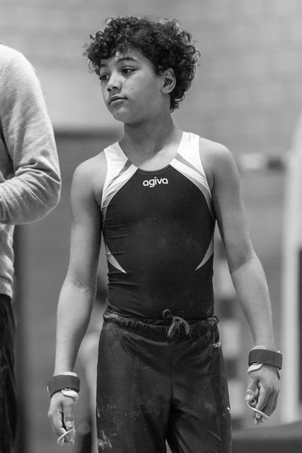 Young gymnast with curly hair stands attentively in Agiva leotard, focused expression showing concentration between routines