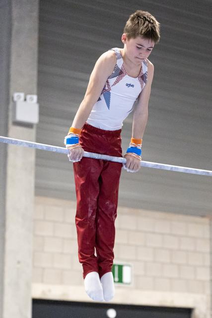 Young gymnast in white and red uniform grips horizontal bar with intense concentration during routine