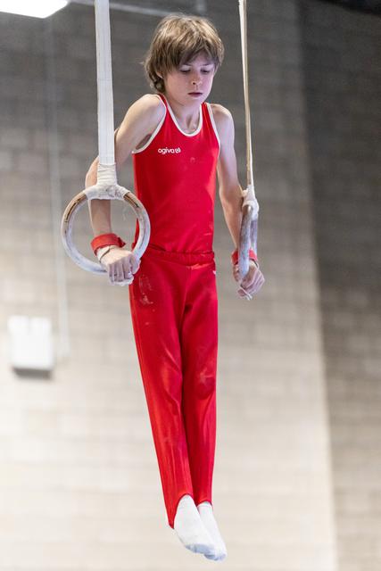 Young gymnast in red uniform performs on rings with chalk-dusted hands, displaying intense concentration during routine