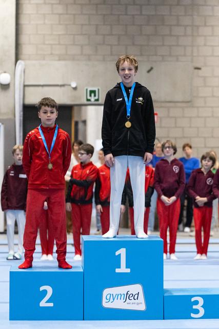 Gold medal winner stands proudly on first place podium wearing black tracksuit and blue medal, with silver medalist beside him