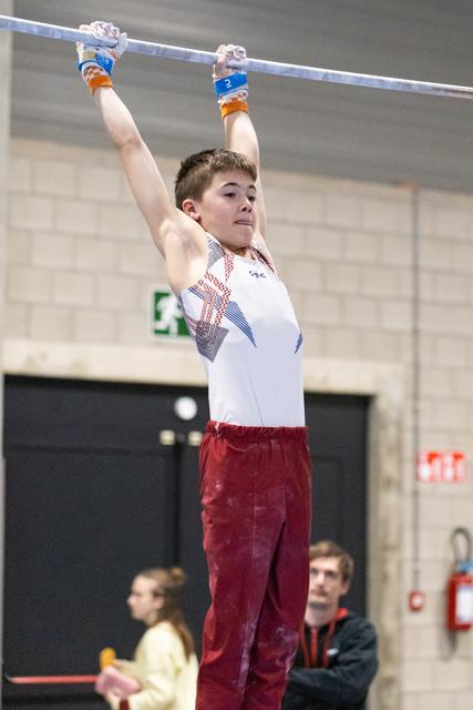 Young gymnast grips the high bar with focused expression, wearing blue wrist guards and red pants during training