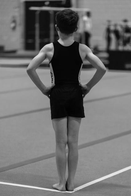 Young gymnast stands with hands on hips, facing away, awaiting her turn on the floor exercise mat in a training facility
