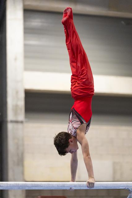 Young gymnast performs a handstand on the balance beam, legs extended straight up, wearing red top and striped shorts