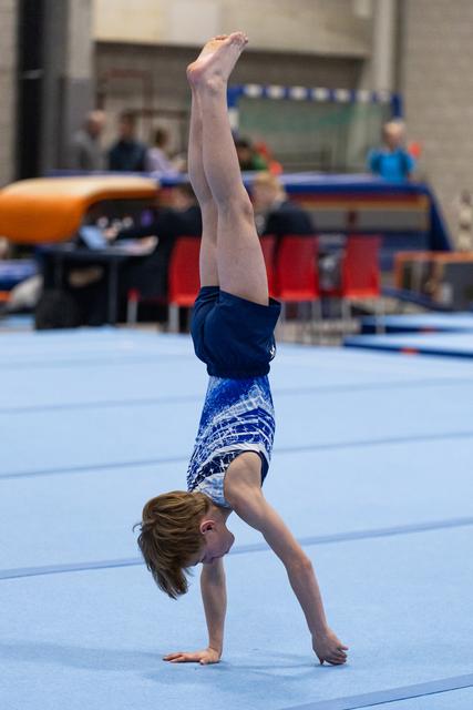 Young gymnast performs a vertical handstand on floor exercise mat, legs straight and pointed upward in blue patterned leotard