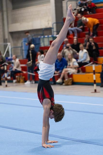 Young male gymnast performs a vertical handstand on competition floor while spectators watch from colorful bleachers