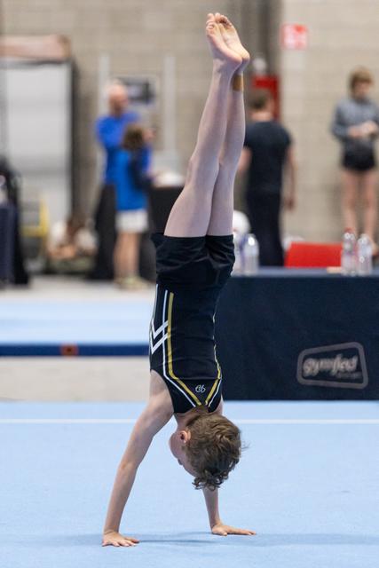 Young gymnast performs a handstand on blue floor mat during routine, legs extended straight up in competitive gym setting