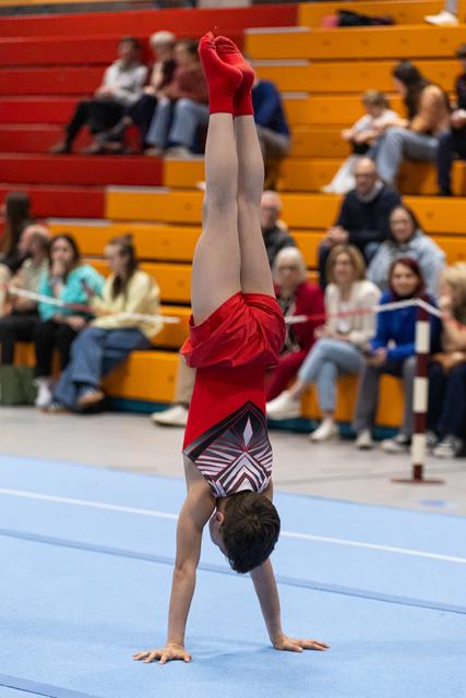 Young gymnast performs a handstand on floor exercise mat wearing red warmup jacket, with spectators in bleachers