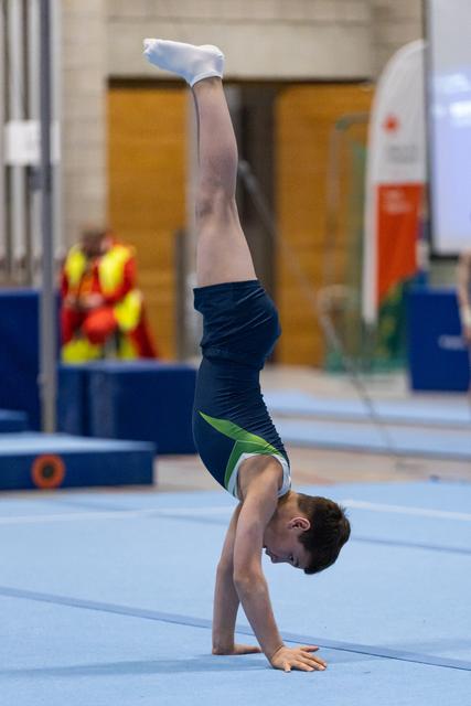 Young gymnast performs a vertical handstand on training mat, legs extended straight up, in indoor gymnastics facility
