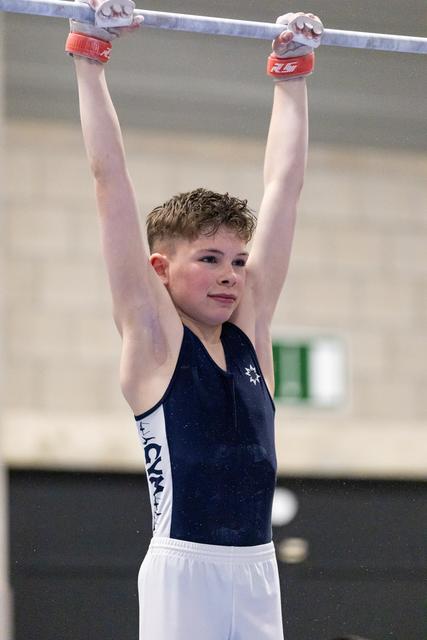 Young gymnast in navy leotard hangs from horizontal bar with red grips, displaying focused determination in training facility