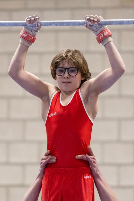 Young gymnast in red leotard and glasses grips horizontal bar while coach supports from below, face showing concentration