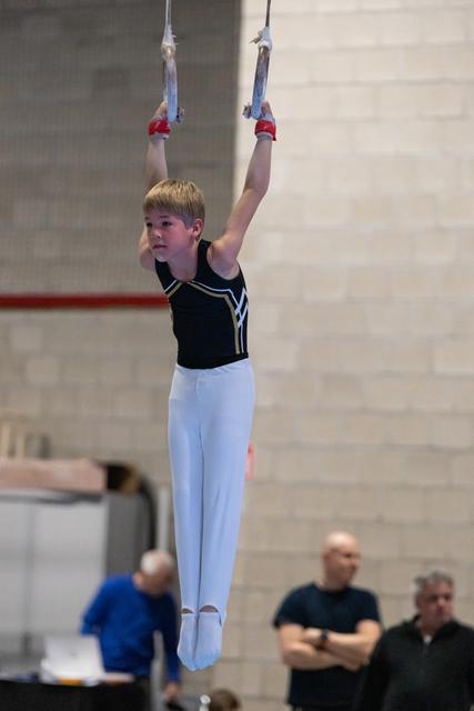 Young gymnast in black and white uniform hangs from rings with focused expression during routine