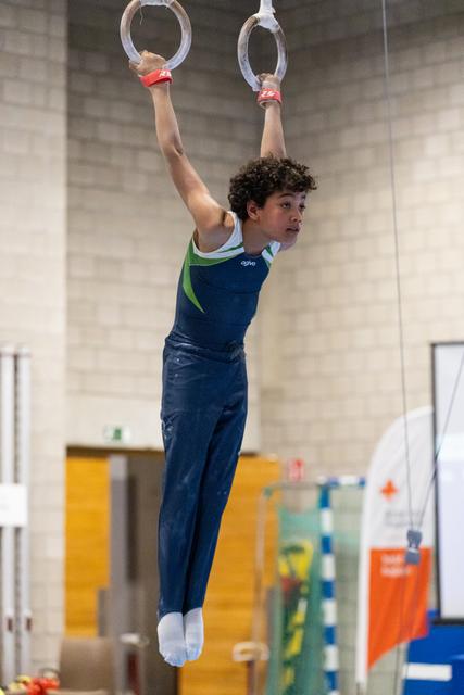 Young male gymnast in navy blue uniform hangs from still rings, demonstrating upper body strength in training facility