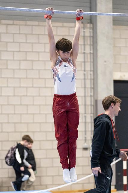 Young gymnast in maroon pants and white leotard hangs from high bar with focused expression during training session
