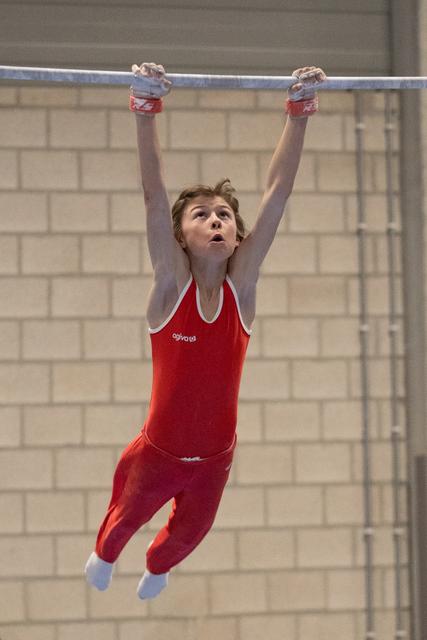 Young gymnast in red leotard hangs from horizontal bar with focused expression, arms extended overhead in training facility