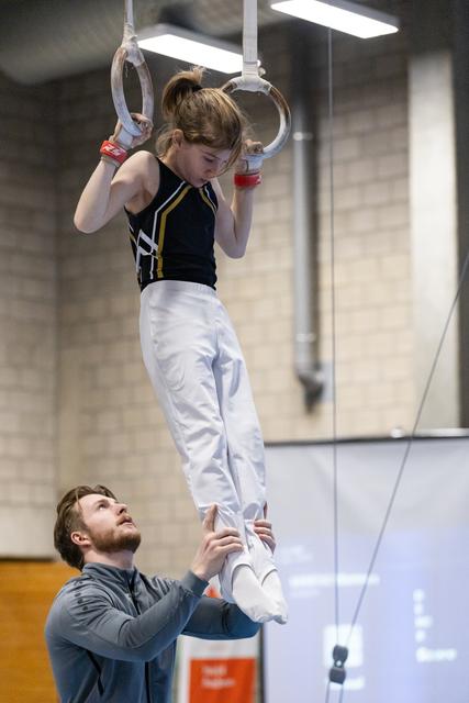 Young gymnast in black and yellow leotard hangs from rings while coach spots from below in indoor training facility
