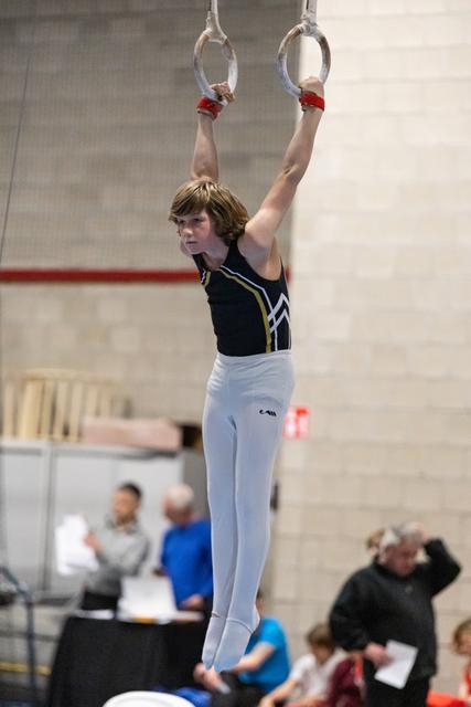 Young male gymnast hangs from rings with determined expression, wearing navy leotard and white pants in gymnasium
