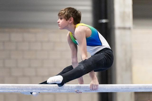 Young gymnast in colorful leotard performs support hold on horizontal bar with focused expression and extended legs