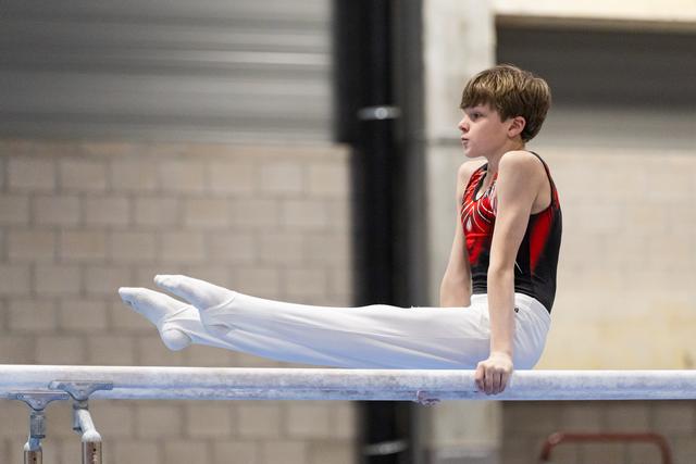 Young male gymnast holds an L-position on horizontal bar, legs extended, focused expression, in indoor gym facility